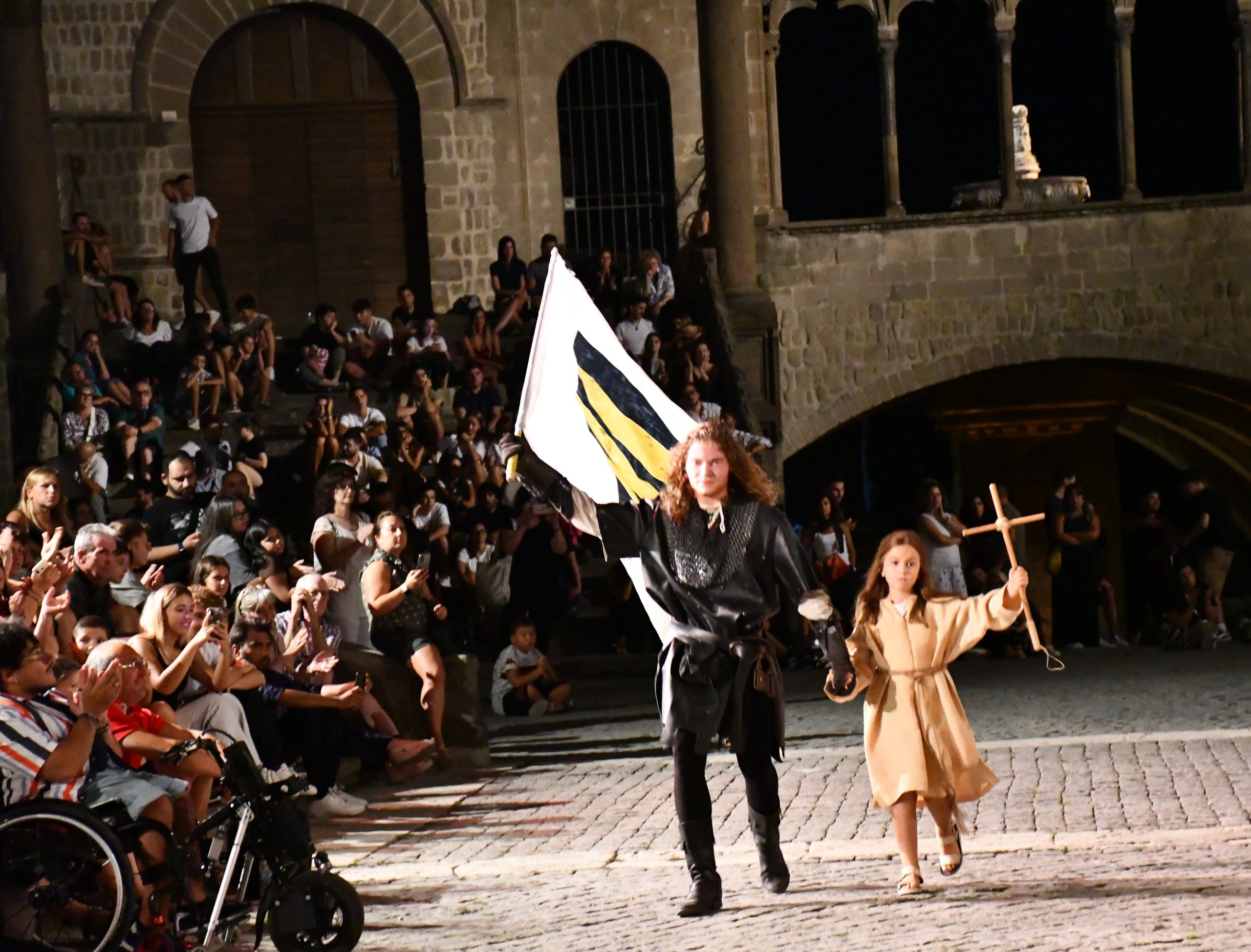 Corteo di rievocazione medievale in piazza con pubblico: un figurante in abiti storici (Capitano del Popolo) porta una bandiera mentre una bambina cammina accanto a lui con una croce (S. Rosa), davanti a spettatori seduti su gradinate.