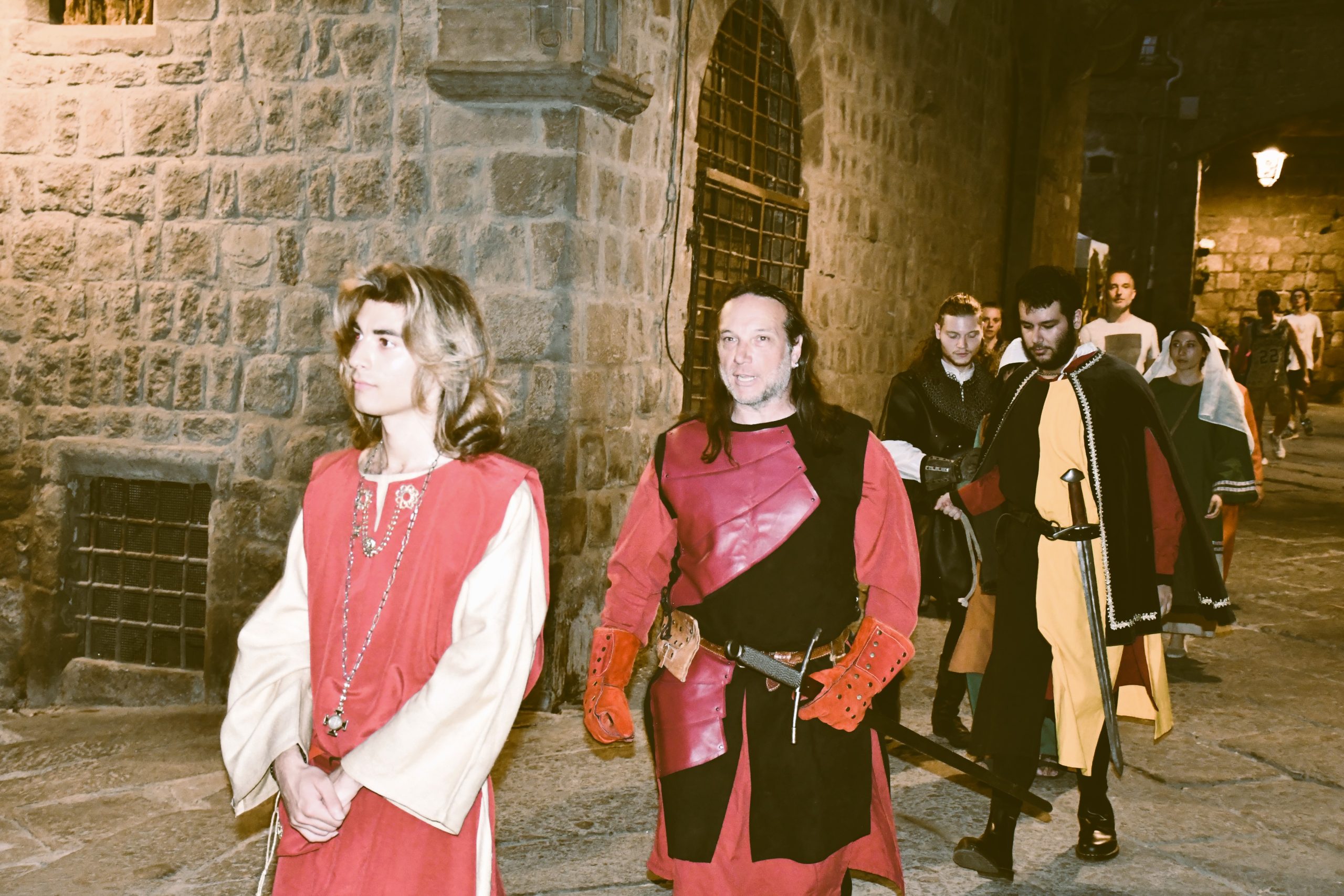Corteo di rievocazione medievale notturna in una strada in pietra: figuranti in abiti storici camminano in fila, guidati da un uomo in armatura rossa e nera e un giovane in tunica rossa.