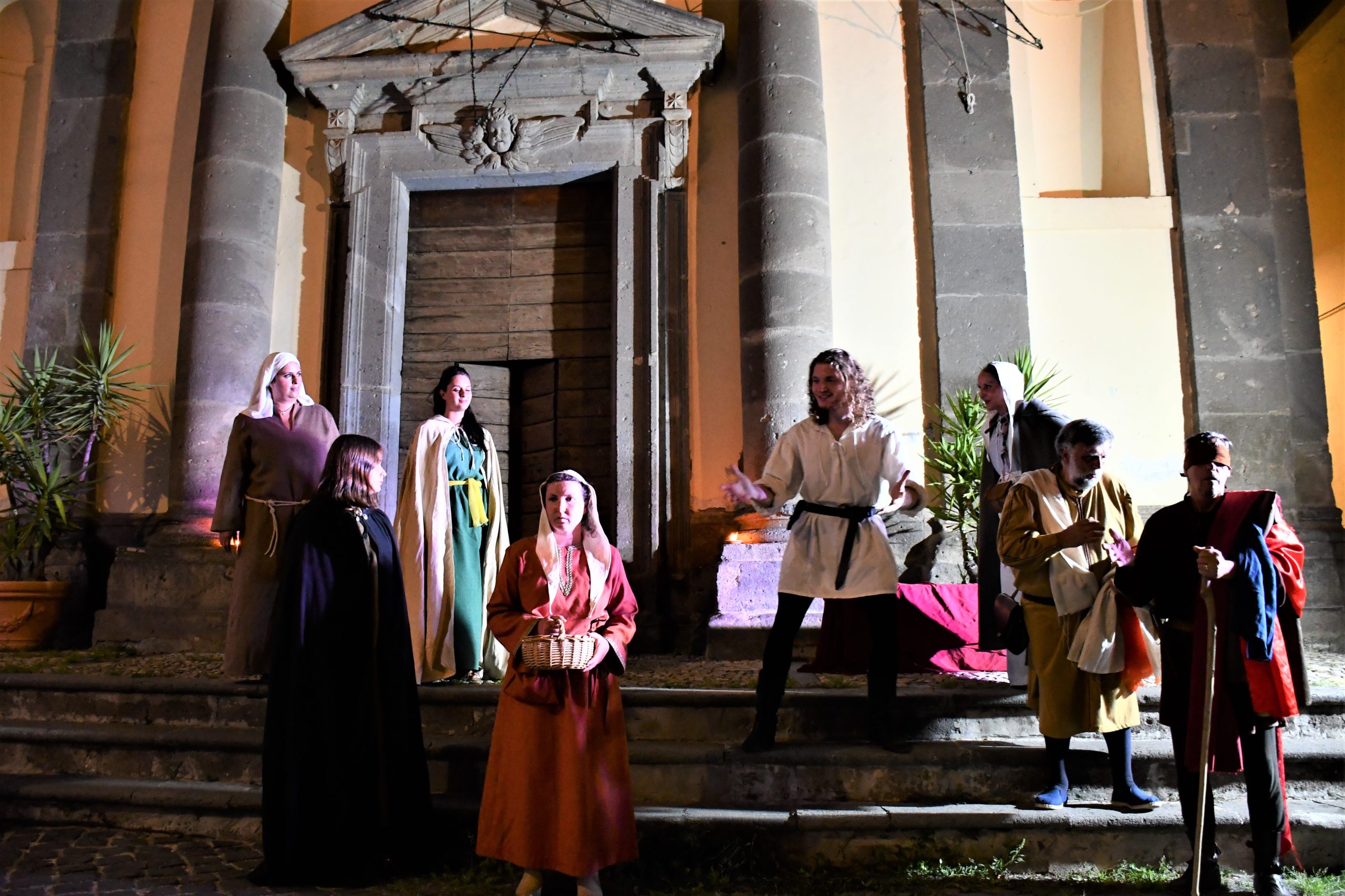 Scena di rievocazione medievale notturna davanti a un edificio storico con colonne: figuranti in abiti d’epoca interpretano una scena teatrale su una scalinata, con una donna in primo piano che tiene un cesto.