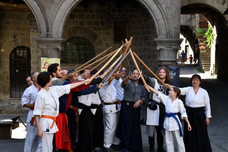Gruppo di figuranti durante una dimostrazione di combattimento storico in un chiostro: partecipanti in abiti da allenamento e costumi medievali incrociano bastoni al centro della scena.