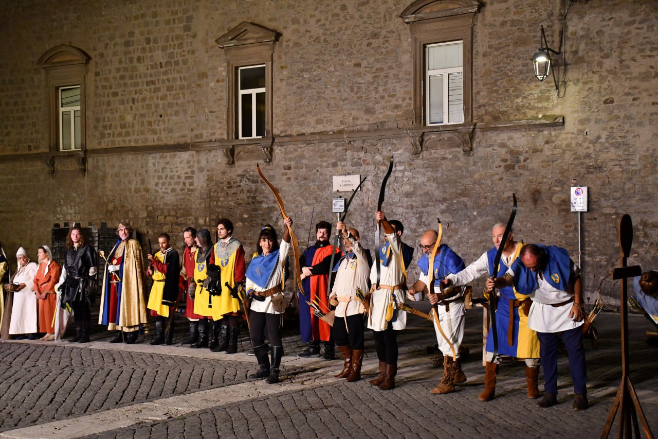 Schieramento di arcieri e figuranti in abiti medievali durante una rievocazione notturna in una piazza storica, con archi alzati e costumi con colori araldici.