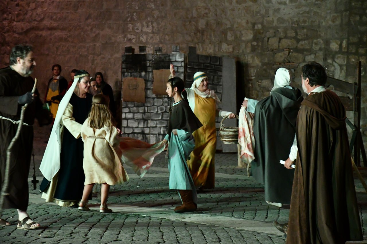 Scena di rievocazione medievale notturna in piazza: donne e bambini in abiti storici partecipano a una rappresentazione, con gesti e movimenti teatrali davanti a una scenografia in pietra.