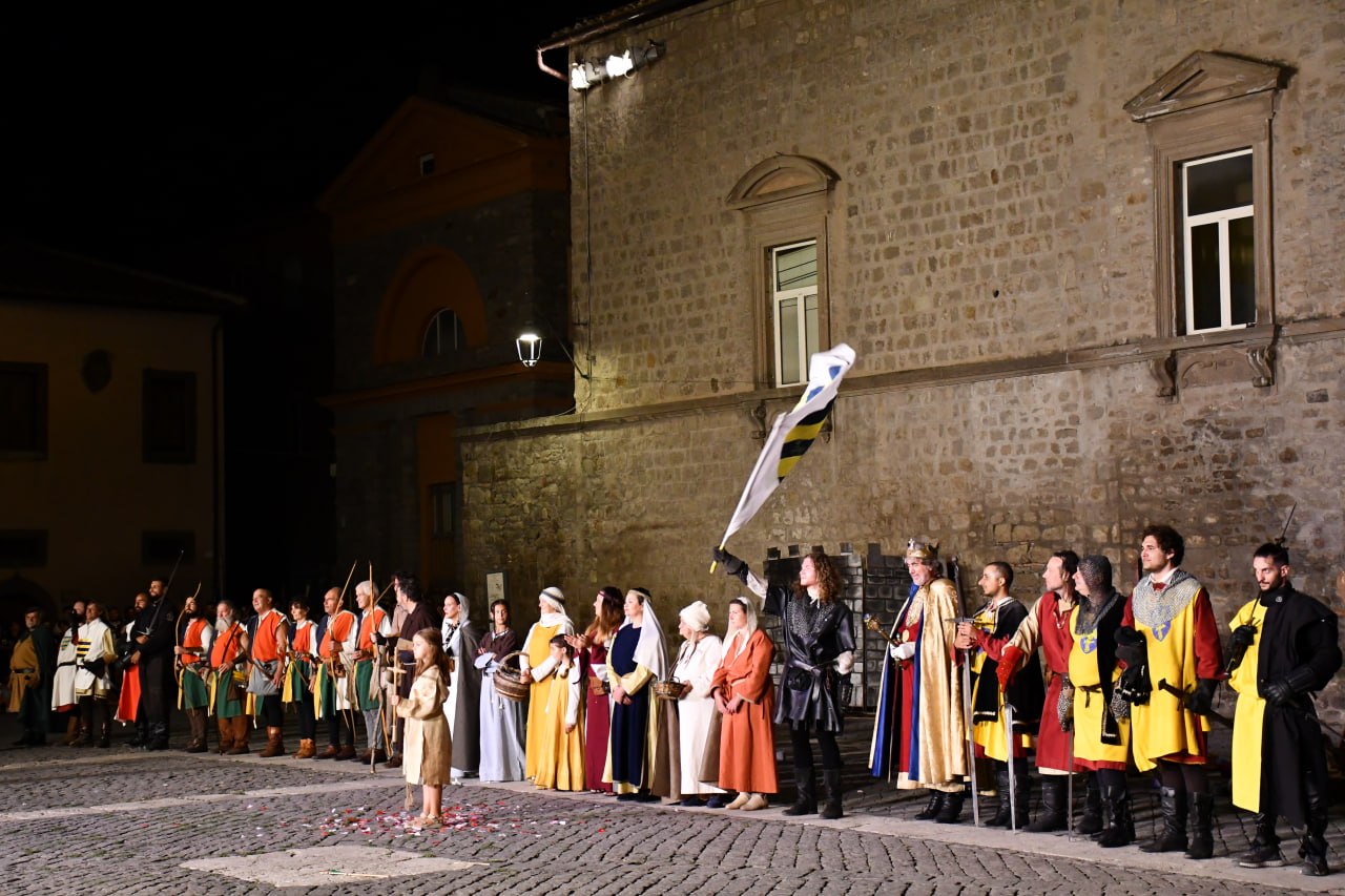 Grande schieramento di figuranti in abiti medievali durante una rievocazione notturna in piazza: uomini, donne e bambini sono disposti in fila mentre uno sventola una bandiera davanti a un edificio in pietra.