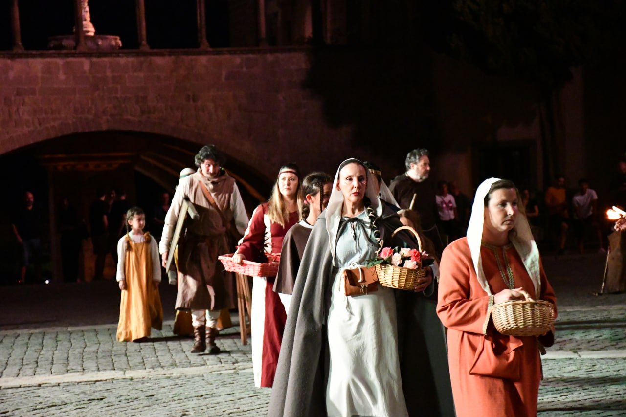 Corteo di rievocazione medievale notturna in piazza: donne e figuranti in abiti d’epoca avanzano portando cesti, seguite da altri personaggi davanti a un edificio storico illuminato.