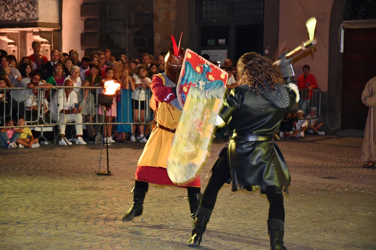 Duello di rievocazione medievale in piazza davanti al pubblico: due figuranti combattono con spade e scudo durante una scena notturna illuminata da torce.