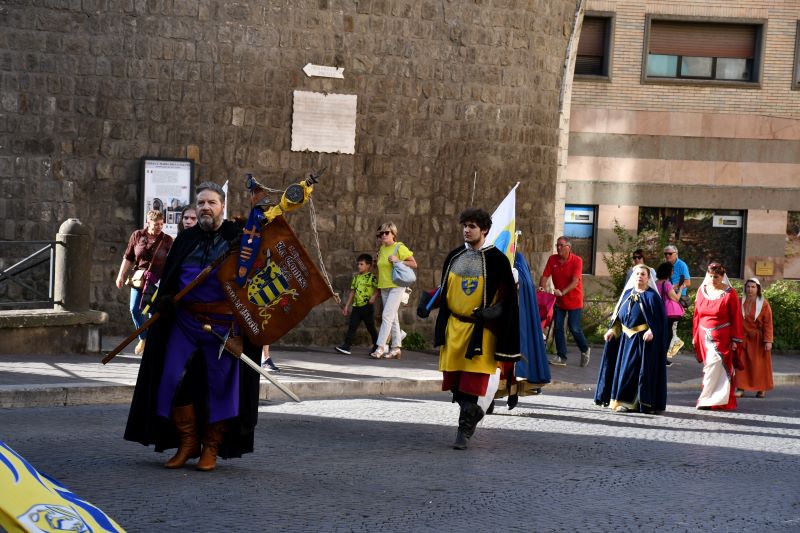Corteo di rievocazione medievale in strada: figuranti in abiti storici sfilano portando stendardi e bandiere, affiancati da passanti in un contesto urbano.